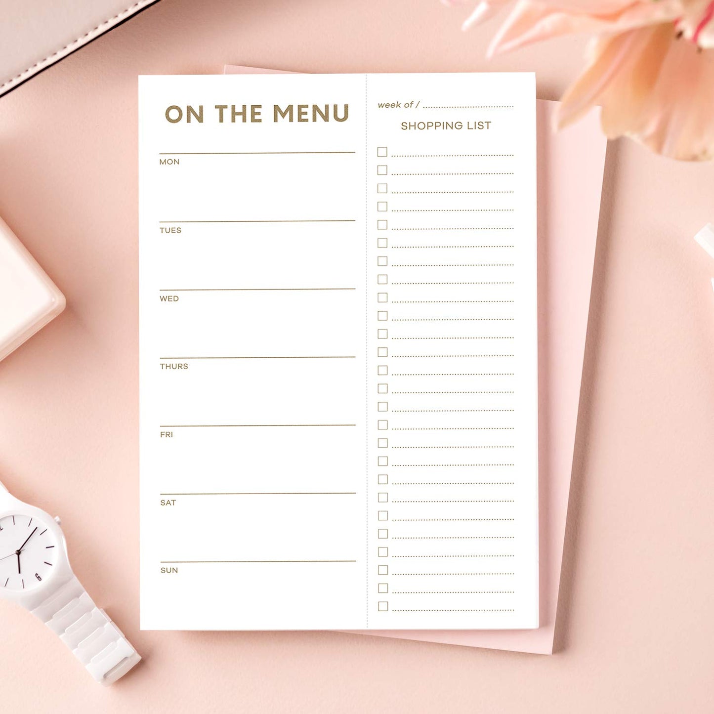 Menu and shopping list planner on a pink surface with a white watch and flowers.