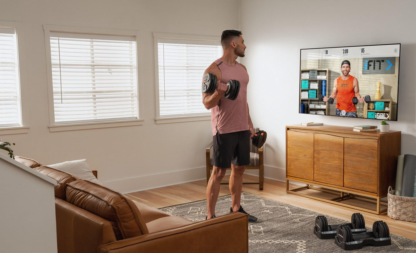 Man exercising with dumbbells in a living room with a TV displaying a fitness program.