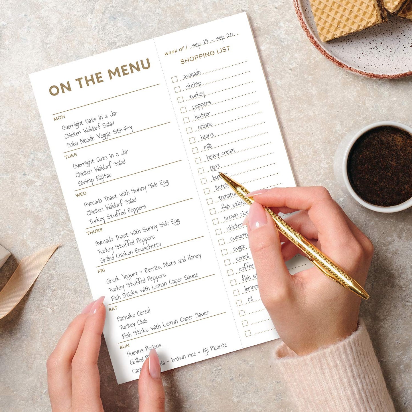Hand holding a pen over a 'On the Menu' and 'Shopping List' sheet on a light surface.