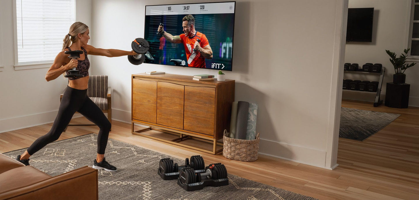 Woman exercising with dumbbells in a living room with a TV displaying a workout video.