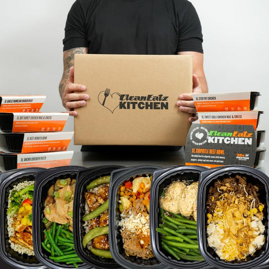 Person holding a Clean Eatz Kitchen box with various prepared meals displayed in front.