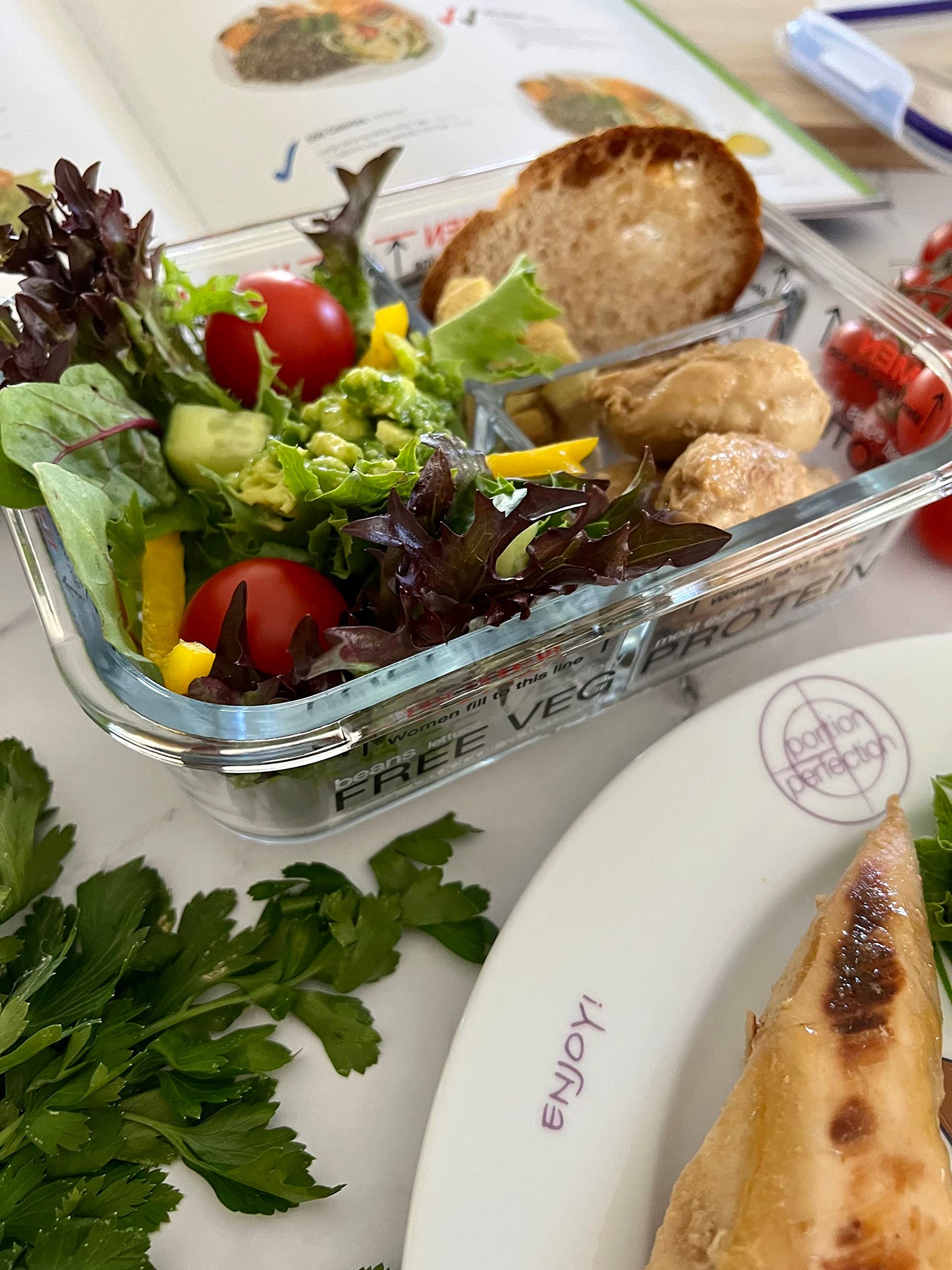 Glass container of salad with bread and crackers on a table with a plate and additional food items.