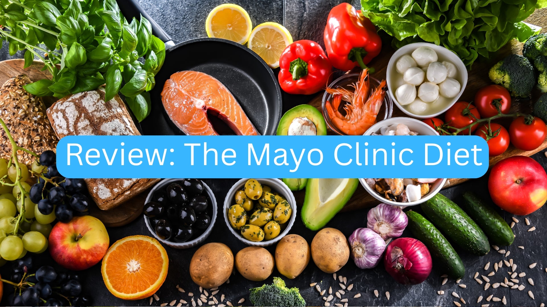 Assorted fruits, vegetables, and a pan of salmon on a dark surface with 'Review: The Mayo Clinic Diet' text overlay.