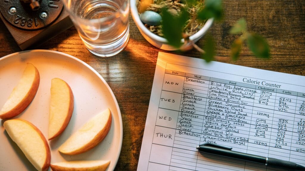 Daily calorie counter and meal tracking for healthy eating. Calorie counter sheet with handwritten meals beside sliced apples and a glass of water