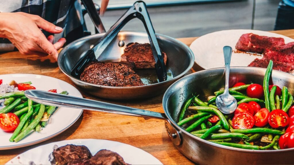 Healthy home-cooked meal with steak, green beans, and tomatoes being prepared in stainless steel pans