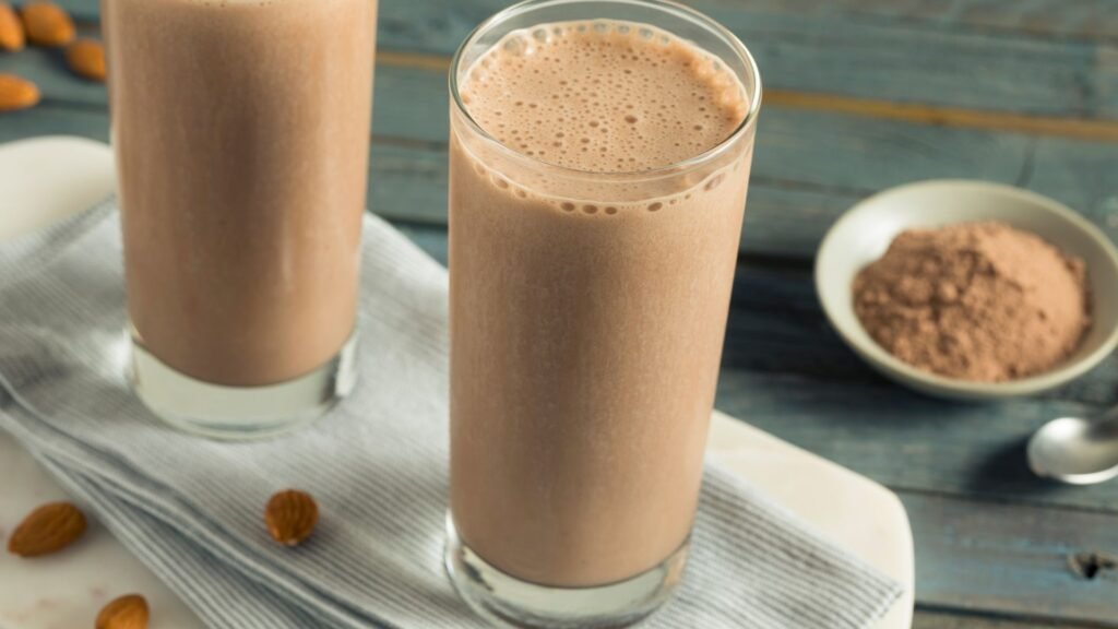 Two glasses of chocolate protein shake with almonds and powder on table